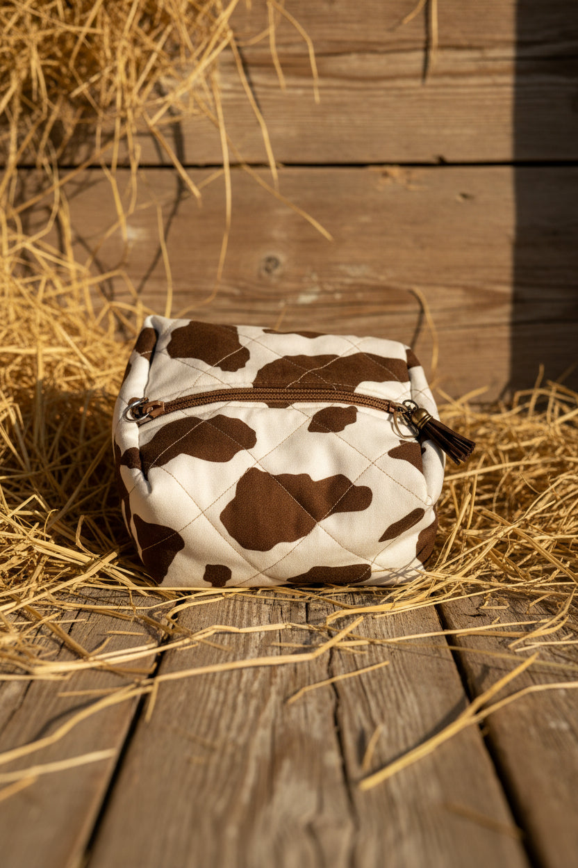 A small zippered pouch with a brown and white cow print pattern, featuring a brown zipper and tassel, resting on a blue surface.