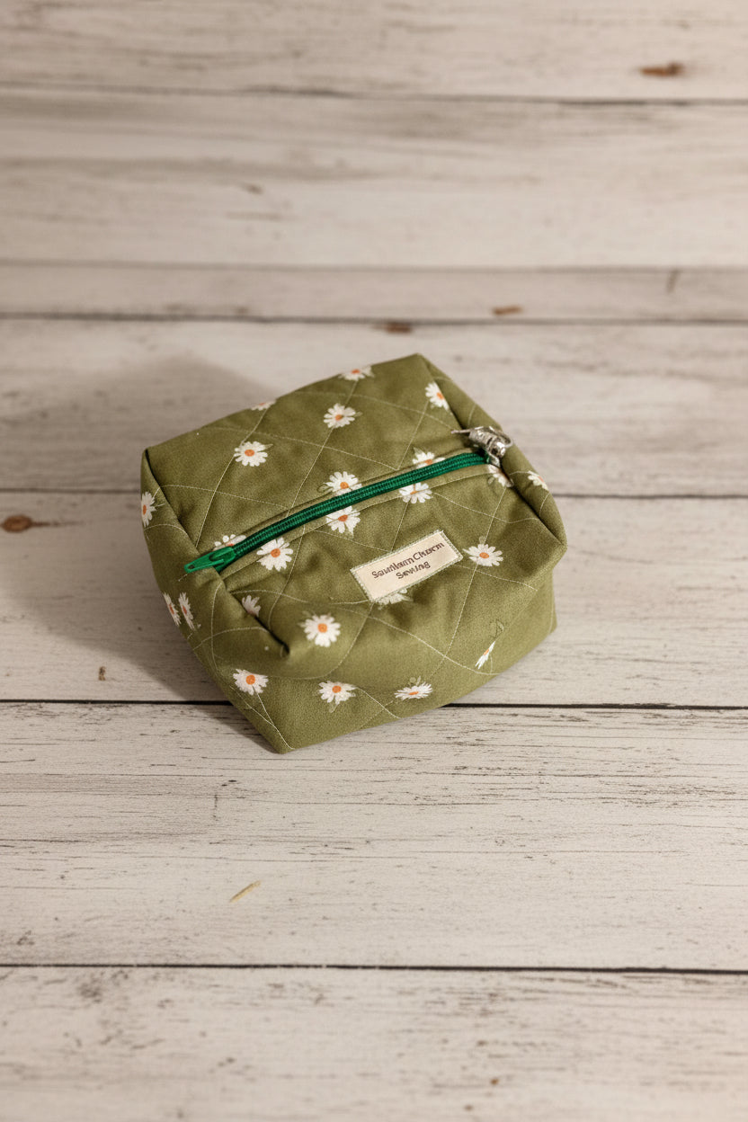An olive green quilted cosmetic pouch with a white daisy floral print and a green zipper, resting on a gray floor next to a matching floral fabric item.