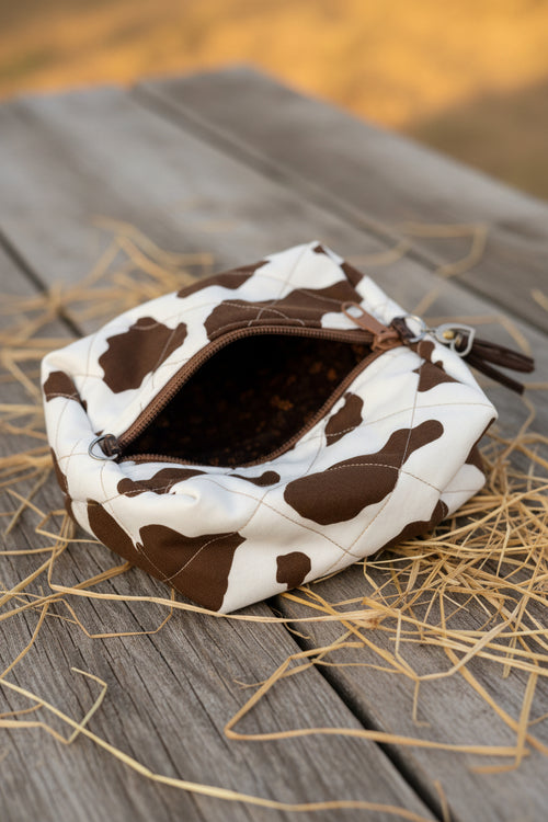 A small quilted pouch with a brown and white cow print pattern, shown open to reveal a brown patterned lining. The pouch has a silver metal zipper and a small silver heart-shaped charm attached near the top right, resting on a blue fabric surface.