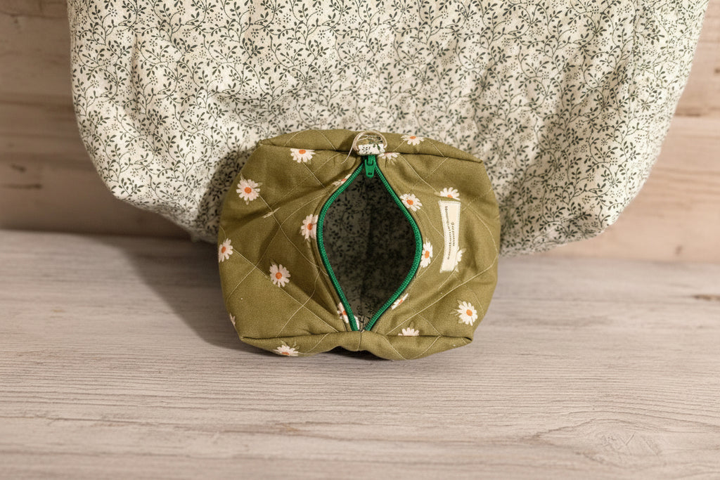 An olive green quilted fabric pouch with a white daisy print and green zipper trim, shown next to a matching quilted textile with a white floral print.