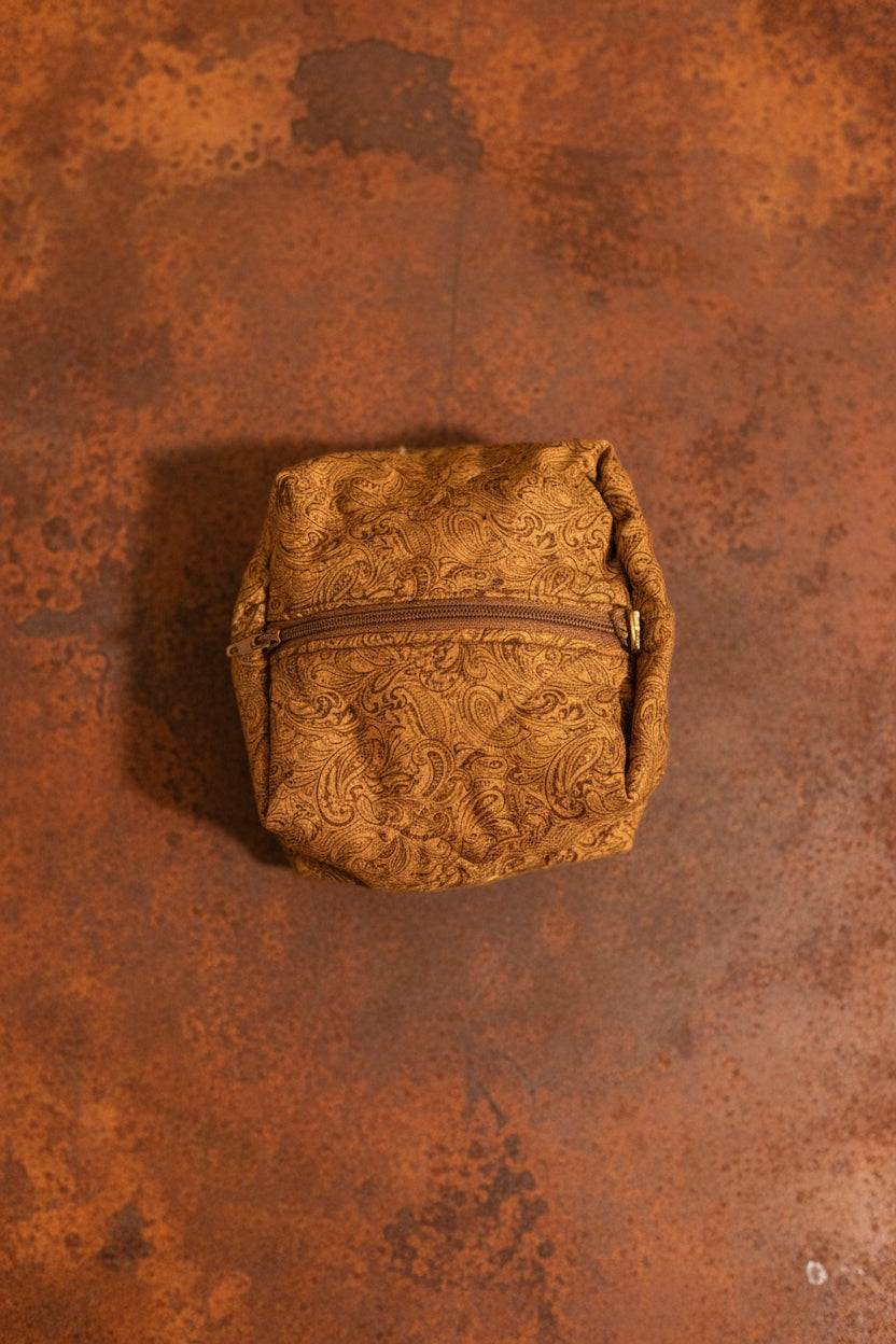 A small textured mustard-brown zippered pouch with a floral jacquard pattern, placed on a light gray surface below a larger matching patterned textile.